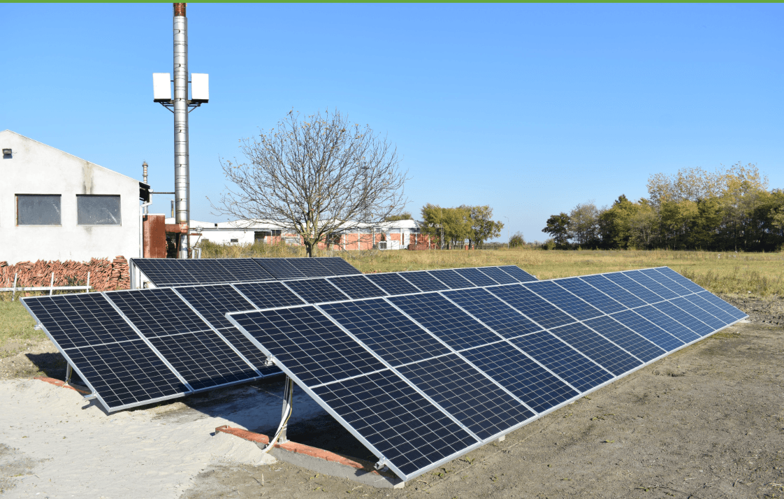Solar panel array in a field with a building and antenna under blue sky.